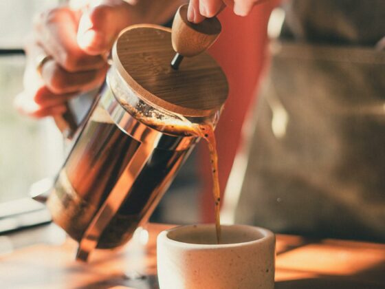 Hands holding French press at 45 degree angle, pouring coffee into small stone cup with light shining through a window in the background.