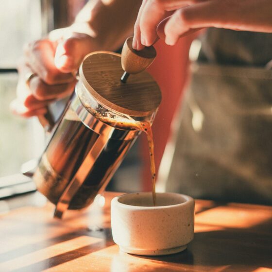 Hands holding French press at 45 degree angle, pouring coffee into small stone cup with light shining through a window in the background.